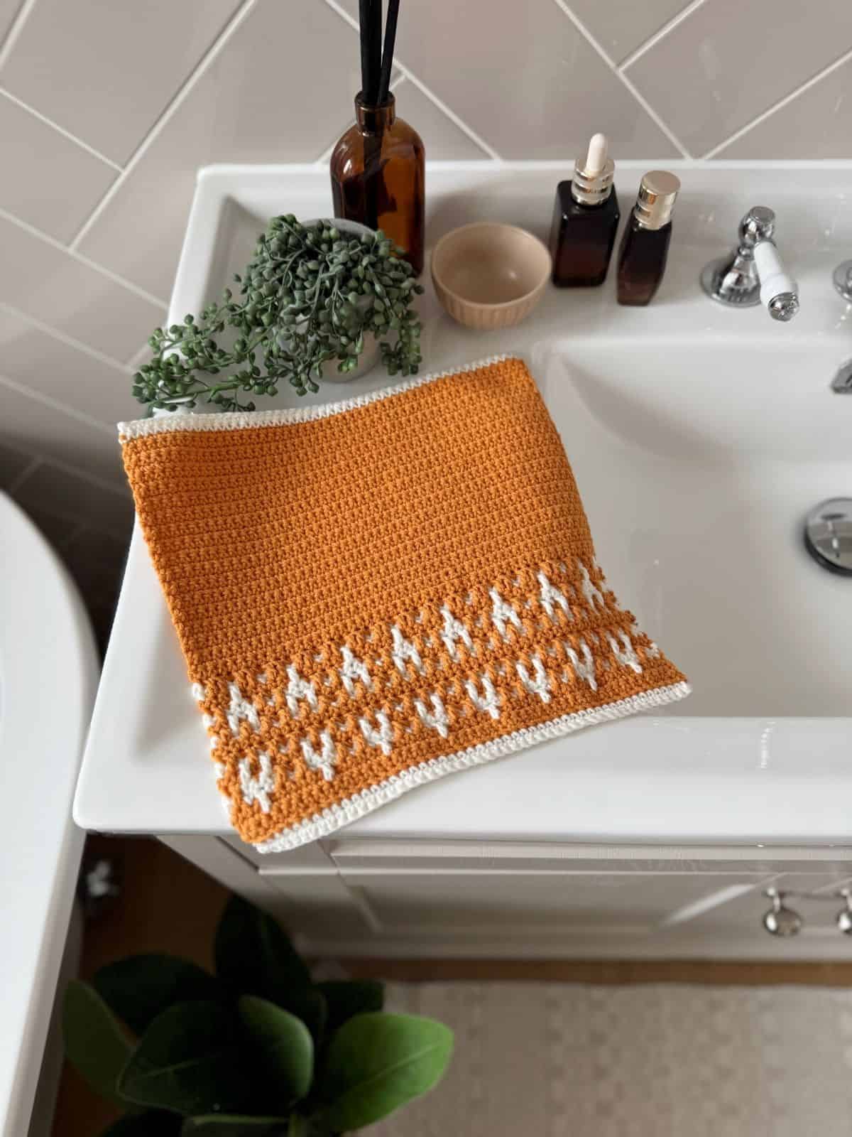 A crocheted orange and white washcloth with a geometric pattern is laid out on a white bathroom sink next to soap bottles, a small bowl, and a plant.