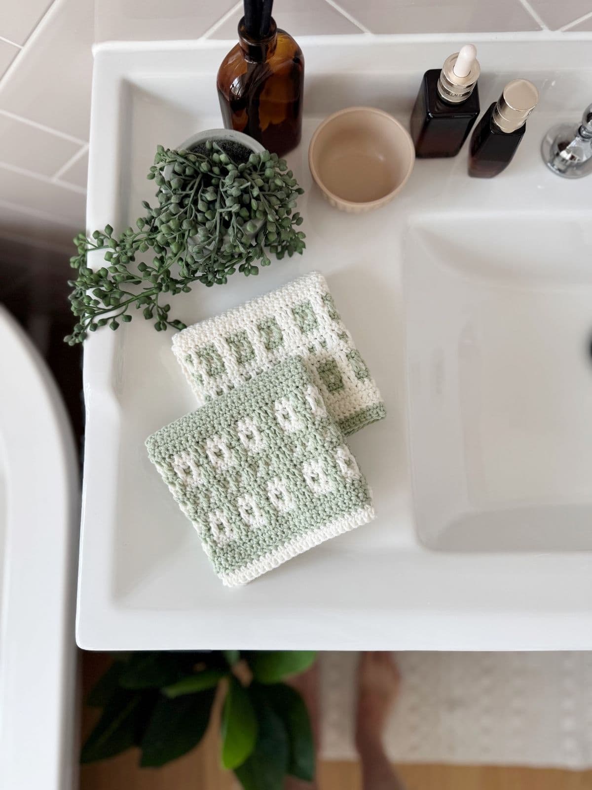 Two crocheted washcloths made with a mosaic crochet free pattern, a small potted plant, brown bottles, and a dish are arranged on a white bathroom sink.