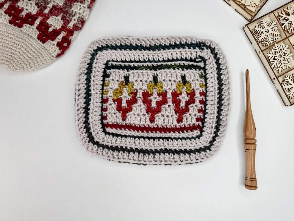 A rectangular crocheted piece with colorful geometric patterns in red, yellow, and black is displayed beside a wooden crochet hook and patterned wooden squares—a perfect setup for a mosaic crochet tutorial on a white surface.