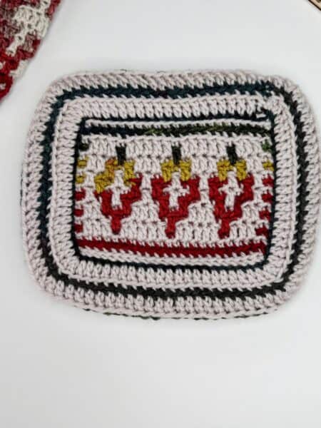 A rectangular crocheted piece with colorful geometric patterns in red, yellow, and black is displayed beside a wooden crochet hook and patterned wooden squares—a perfect setup for a mosaic crochet tutorial on a white surface.