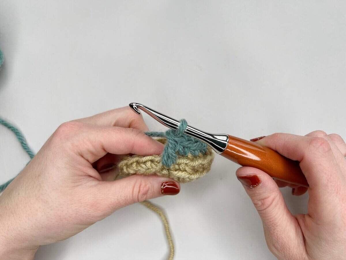 Two hands crocheting with beige and teal yarn using a wooden-handled crochet hook against a white background, perfect for illustrating a mosaic crochet tutorial.