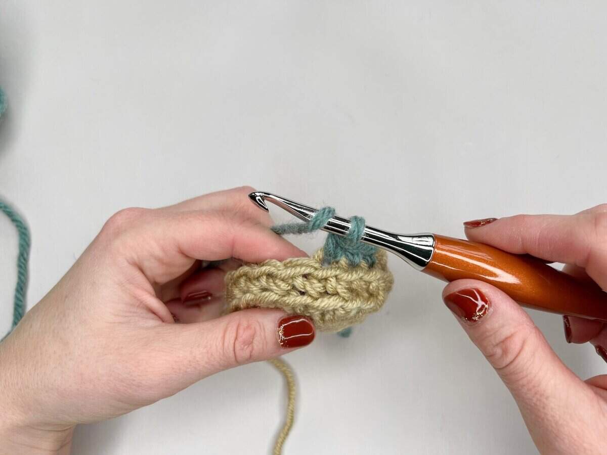Two hands crochet with a wooden-handled hook and beige and teal yarn against a white background, creating a stunning piece perfect for any mosaic crochet tutorial.