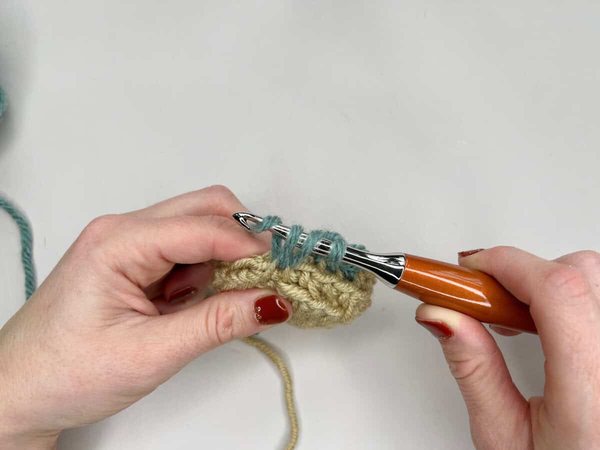 A person follows a mosaic crochet tutorial, using a wooden hook with blue yarn to stitch into beige crocheted fabric, with both hands visible against a white background.