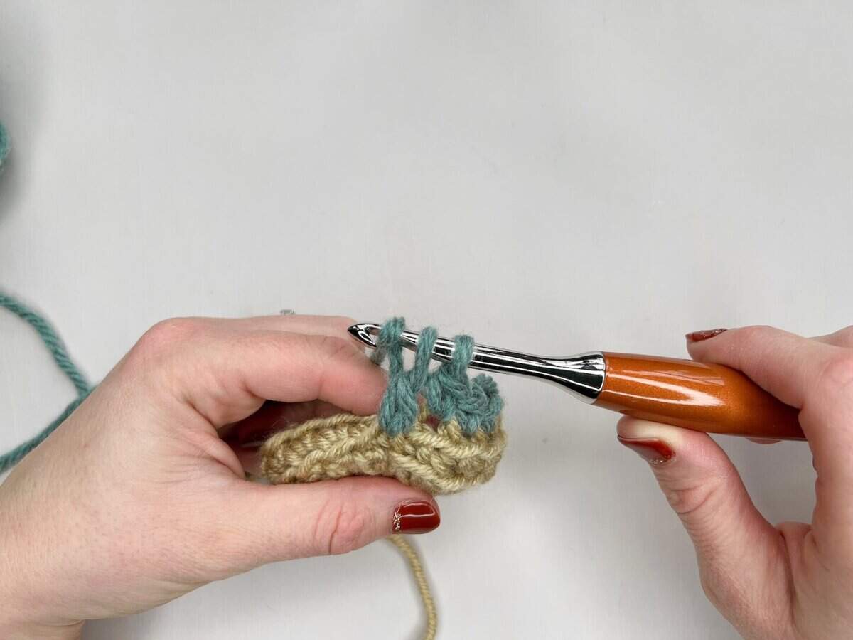 Two hands are crocheting with beige and teal yarn, using a wooden-handled crochet hook to work stitches into the yarn in this mosaic crochet tutorial.