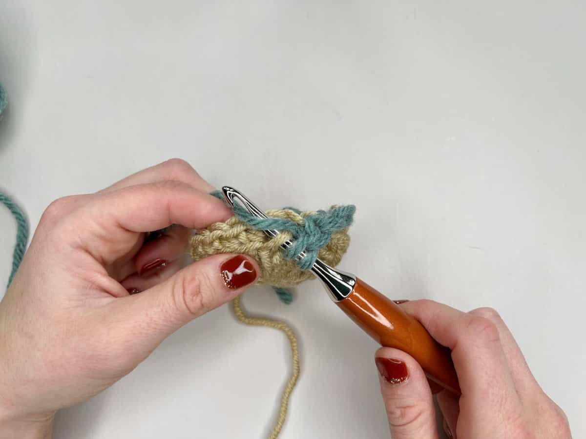 Two hands hold a piece of crochet work, using a wooden-handled crochet hook to stitch blue yarn into beige yarn against a white background, perfect for following a mosaic crochet tutorial.