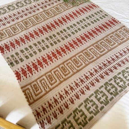 A mosaic crochet blanket pattern with geometric designs in green, red, and beige tones is laid out on a bed, complemented by a small potted plant at the head.