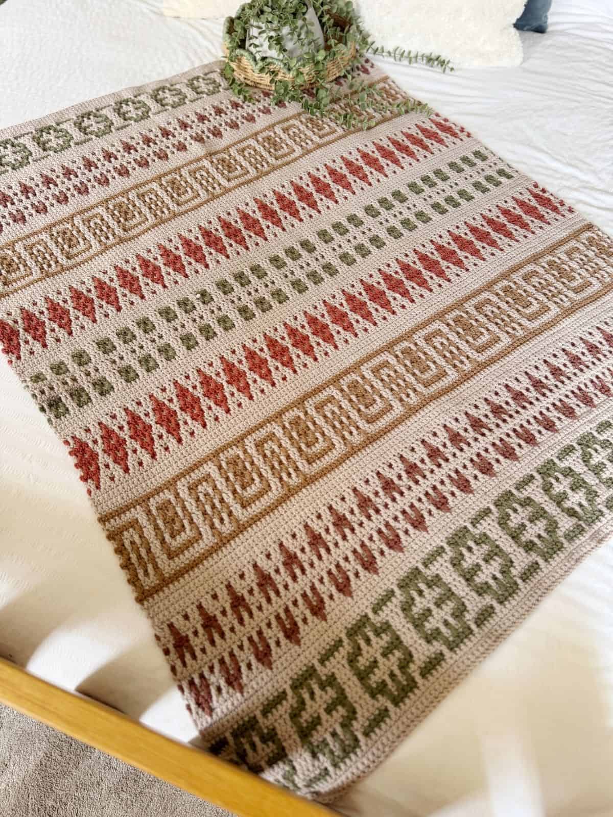 A mosaic crochet blanket pattern with geometric designs in green, red, and beige tones is laid out on a bed, complemented by a small potted plant at the head.