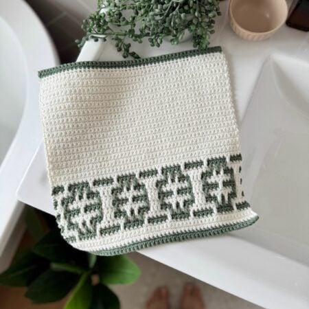 A white crocheted towel featuring an easy mosaic crochet pattern with a green geometric border is draped over a white sink; a plant, brown bottle, and beige cup are nearby.