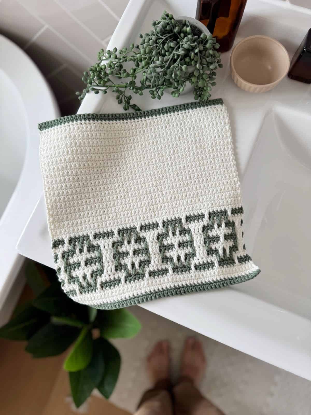 A white crocheted towel featuring an easy mosaic crochet pattern with a green geometric border is draped over a white sink; a plant, brown bottle, and beige cup are nearby.