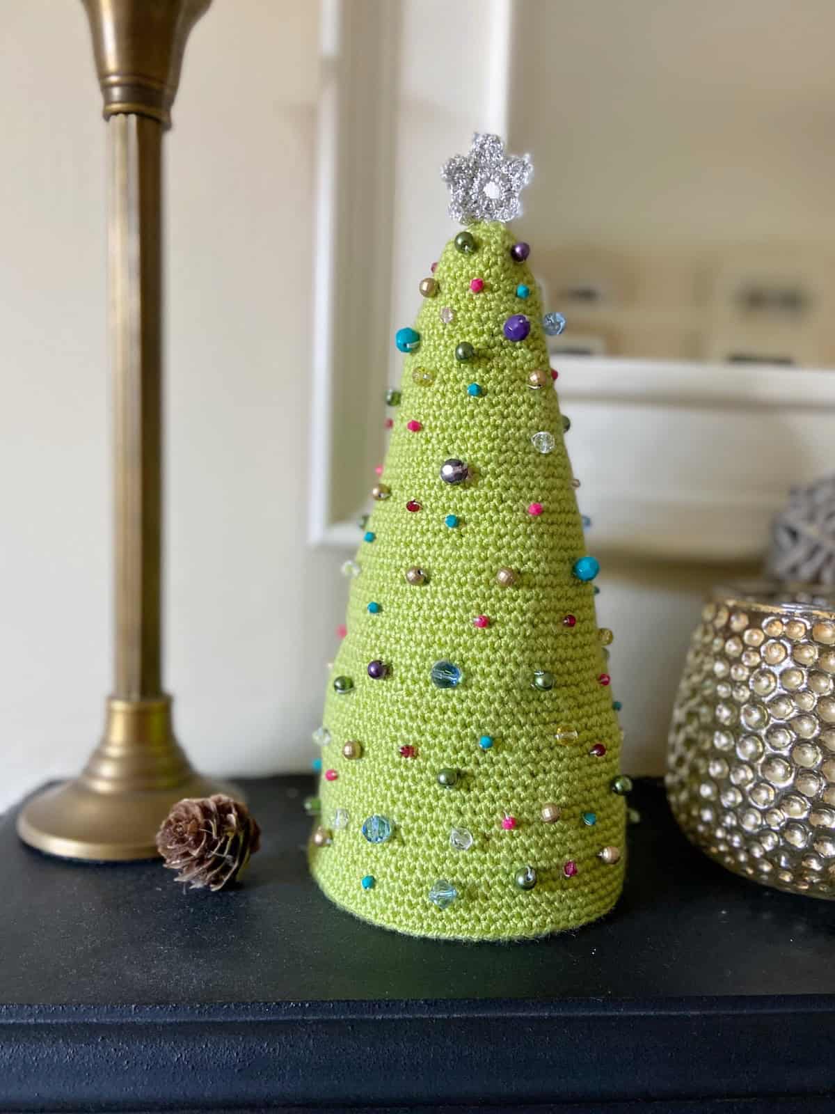 A crochet green cone-shaped Christmas tree, inspired by easy Christmas crochet patterns, with multicolored beads and a silver star topper sits on a black surface next to a gold decorative item and a pinecone.
