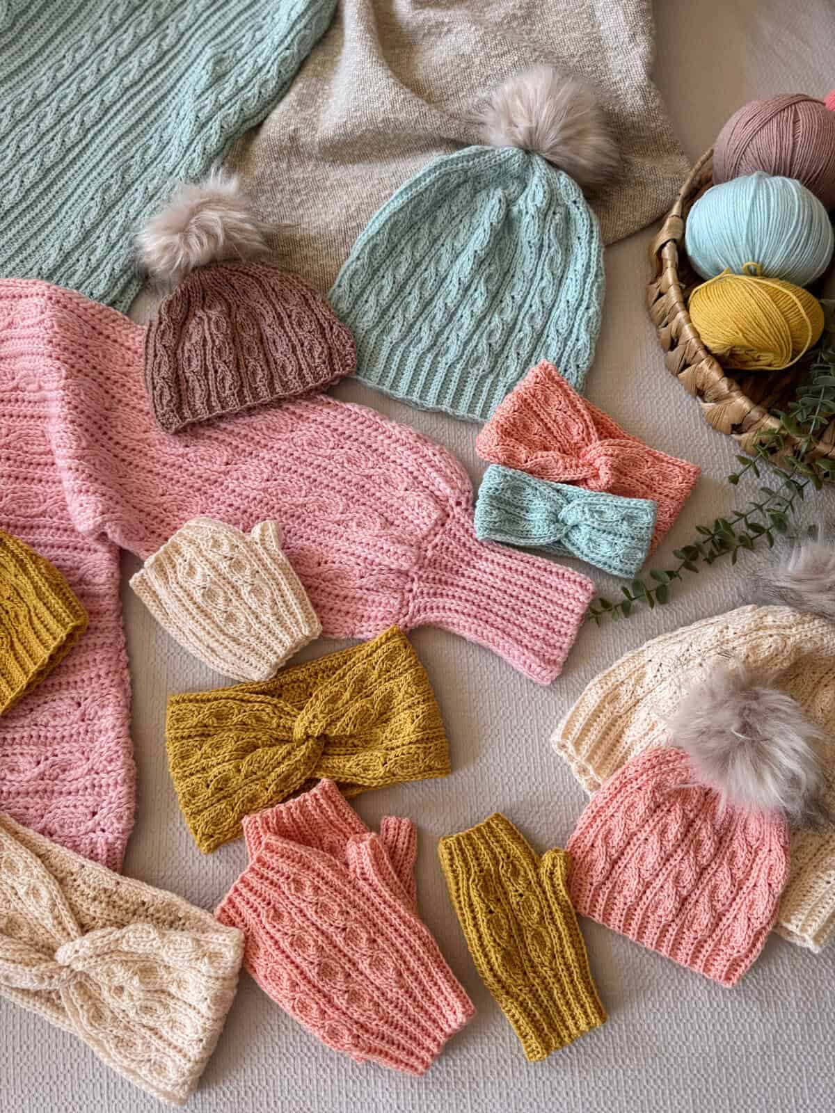 Various knitted hats, headbands, mittens, and sweaters in pastel colors are arranged on a surface, with balls of yarn and a small sprig of greenery to the side.