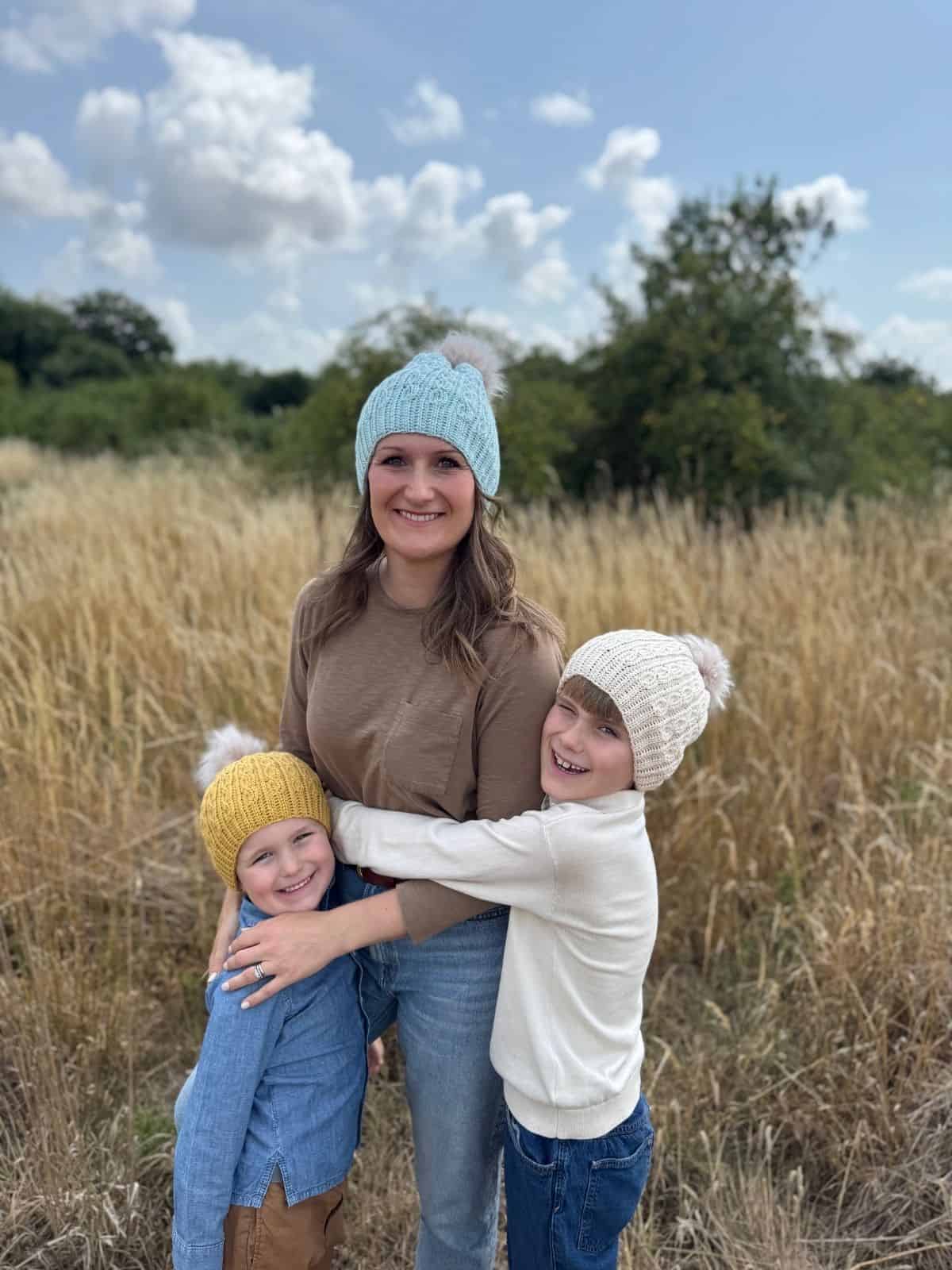 A woman and two children wearing crochet hats, made with a short row crochet hat pattern, stand together in a grassy field, smiling and hugging, with trees and a cloudy sky in the background.