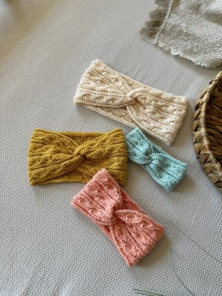 Four headbands in cream, mustard yellow, light blue, and pink—each featuring a twisted crochet headband pattern—are arranged on a light textured surface beside a woven basket and a fringed cloth.