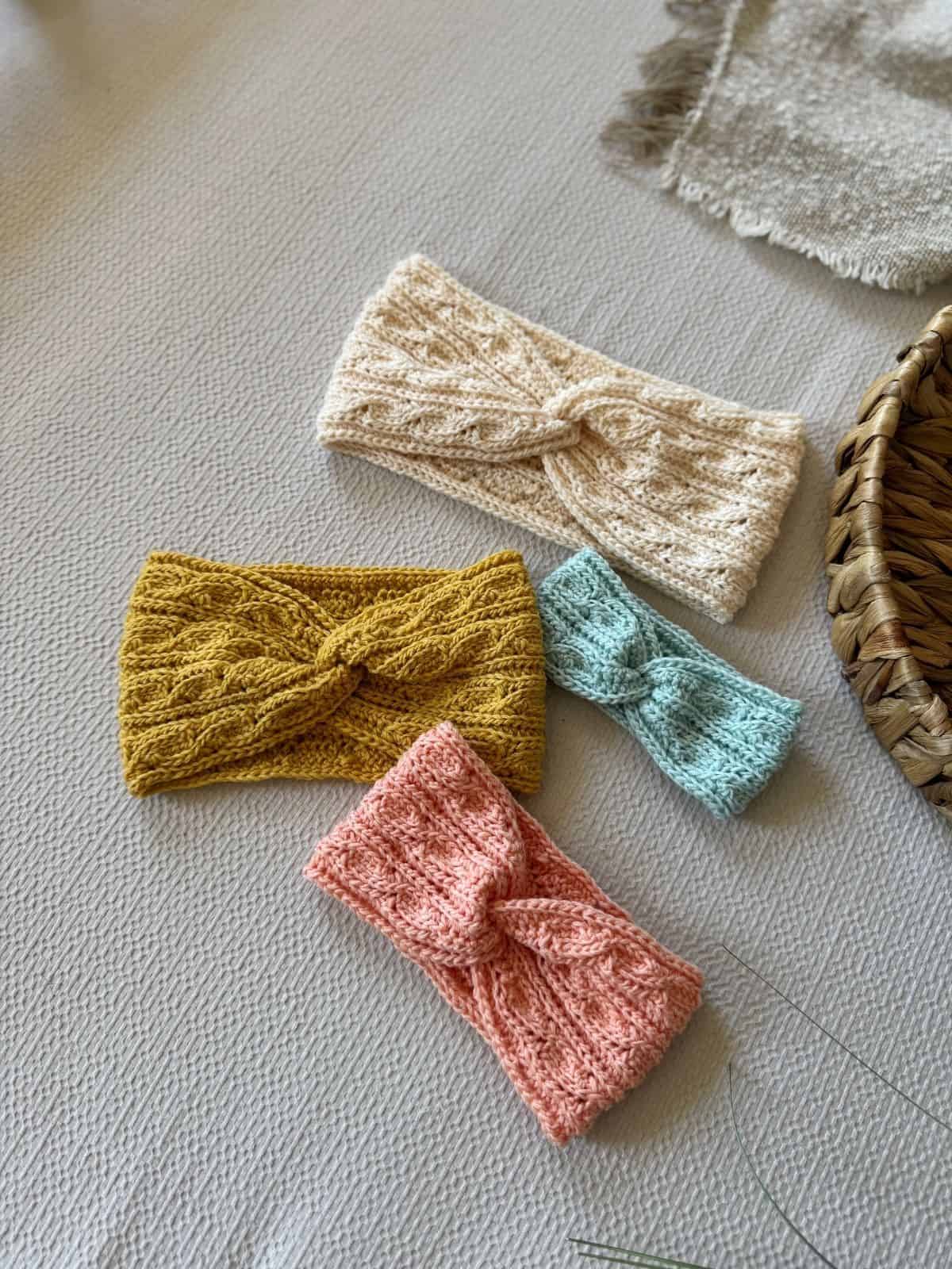 Four headbands in cream, mustard yellow, light blue, and pink—each featuring a twisted crochet headband pattern—are arranged on a light textured surface beside a woven basket and a fringed cloth.