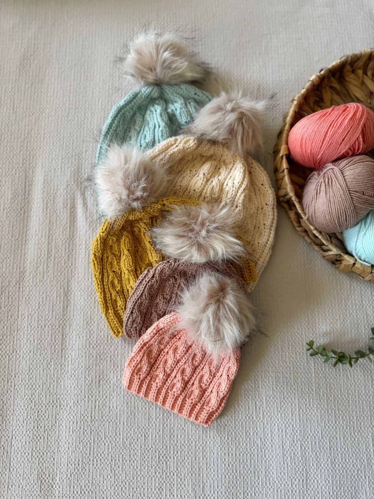 Five crocheted hats with faux fur pom-poms in pastel colors are arranged on a textured surface next to a basket of colorful yarn balls, perfect inspiration for your next short row crochet hat pattern.