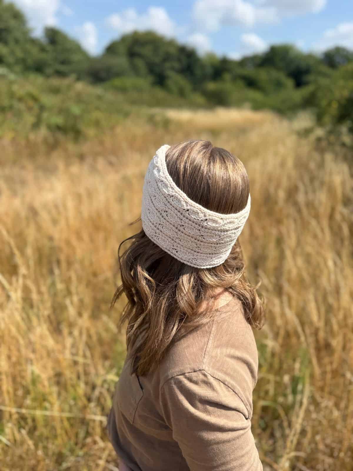 A person with wavy brown hair, wearing a tan top and a twisted crochet headband pattern in beige, stands in a field of tall, dry grass facing away from the camera. Trees and blue sky are visible in the background.