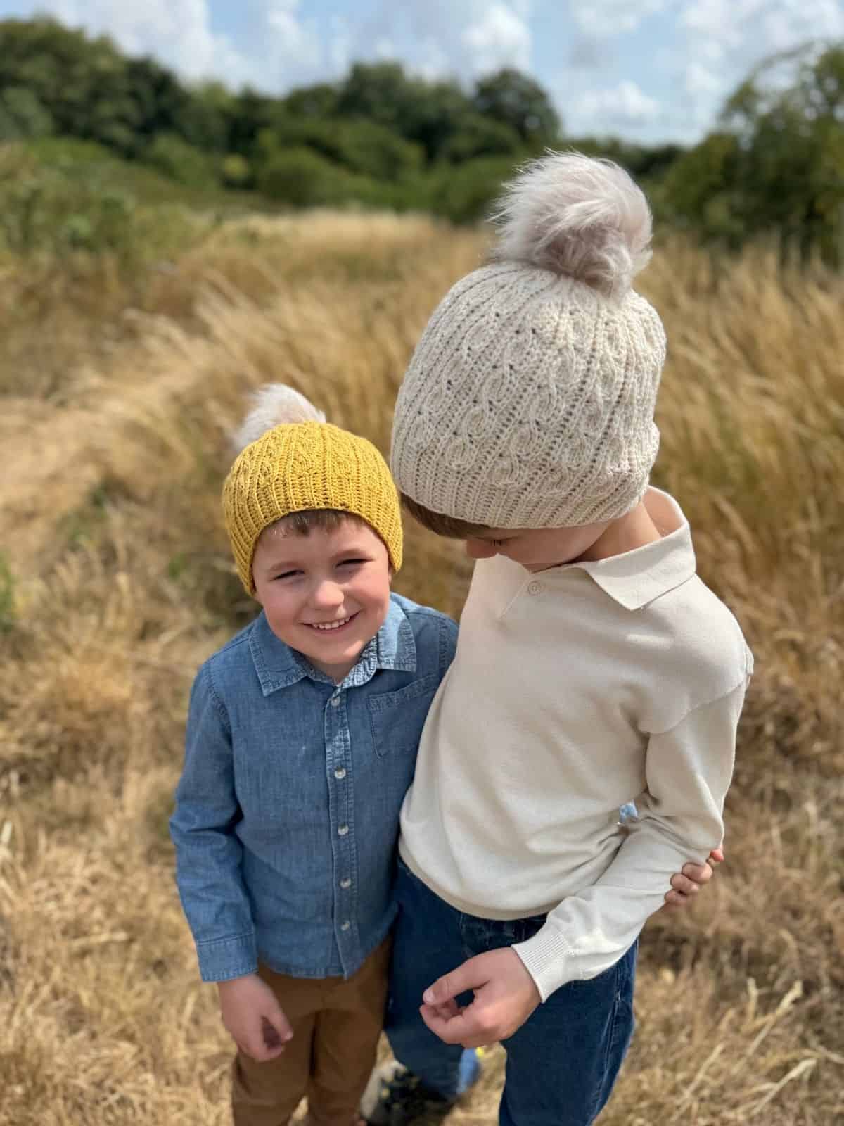 Two children stand on a grassy path outdoors. The younger smiles at the camera in a yellow short row crochet hat, while the older looks down, wearing a cream crochet hat and white sweater.