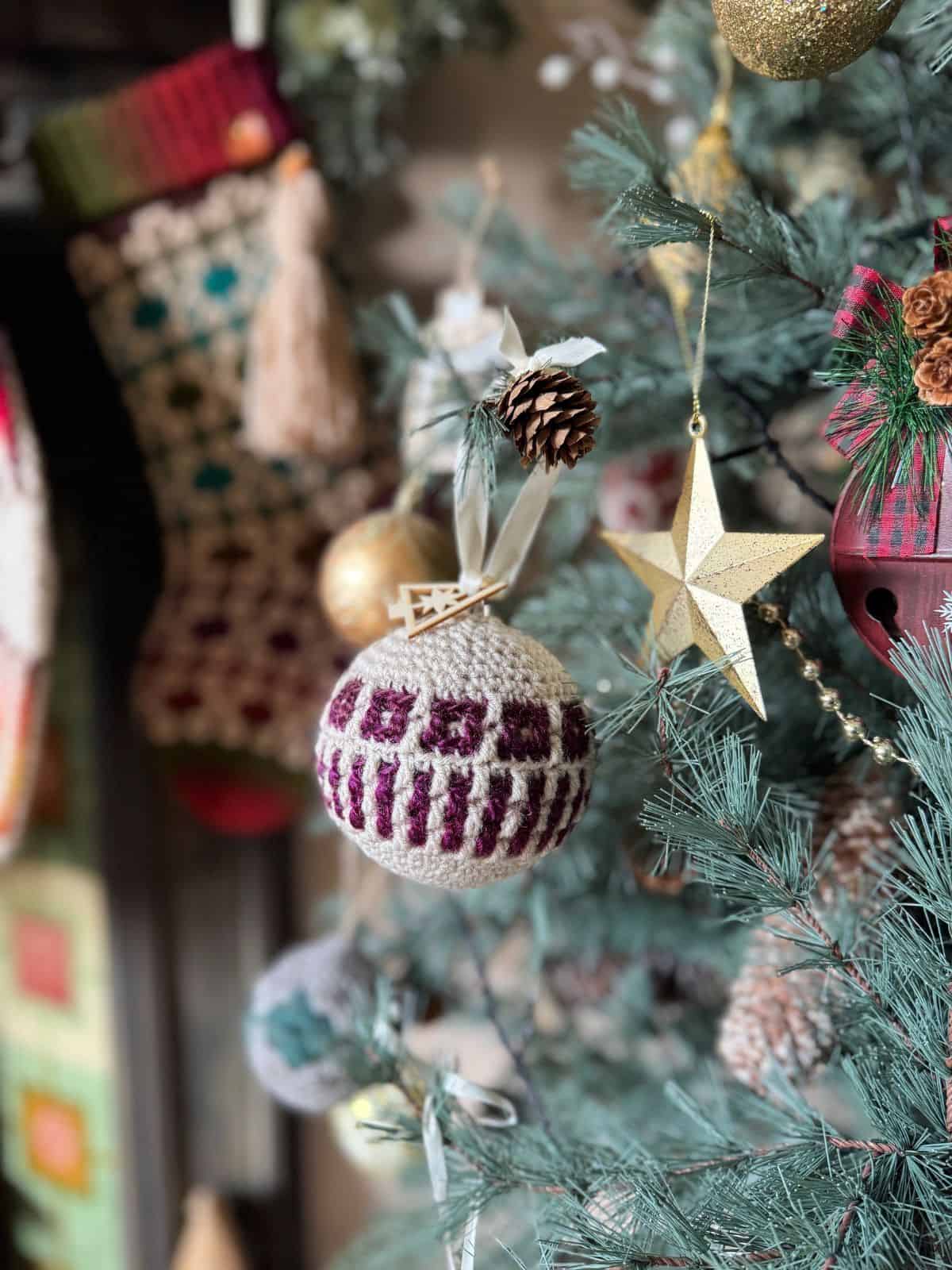 A close-up of a Christmas tree decorated with a knitted ornament, gold star, pine cone, and a stocking in the background—perfect inspiration for easy Christmas crochet patterns.