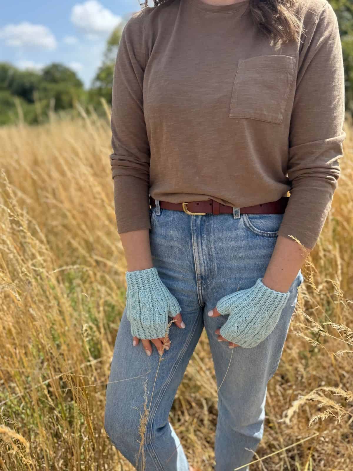 Person standing in a field wearing a brown long-sleeve shirt, blue jeans, a brown belt, and light blue fingerless crochet gloves.