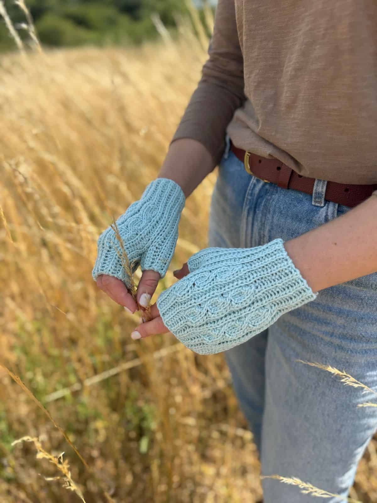 Person standing in a field wearing light blue crochet fingerless mittens pattern free gloves, a brown belt, tan top, and blue jeans, holding a piece of grass.