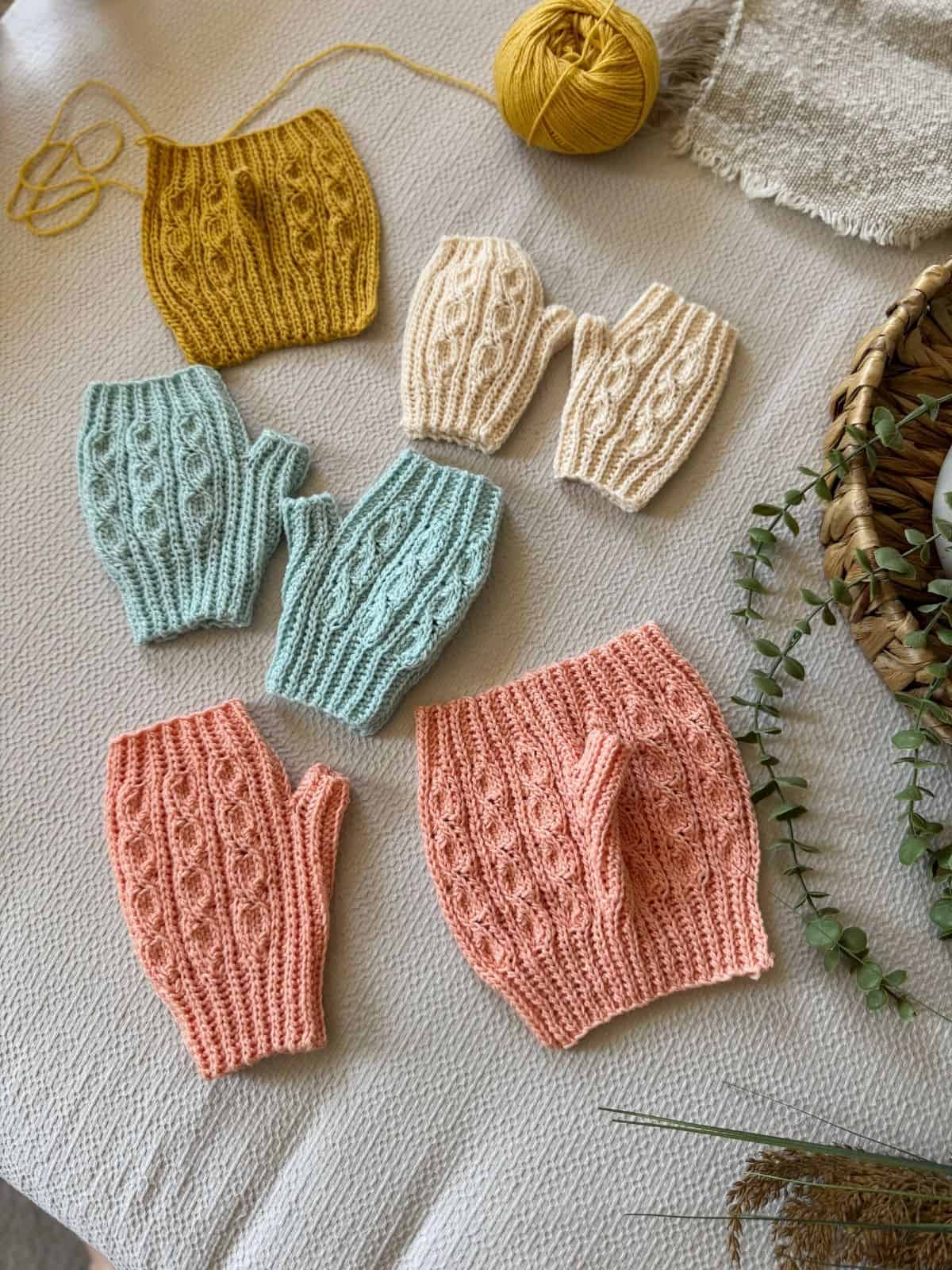 Colorful crochet fingerless gloves and wrist warmers in yellow, cream, blue, and pink are arranged on a table with yarn and a basket—perfect inspiration if you're searching for a crochet fingerless mittens pattern free of charge.