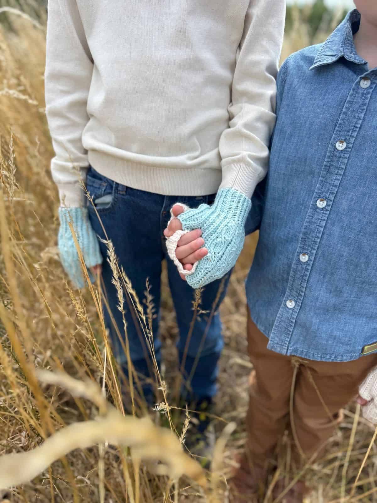 Two people stand in tall grass, one in a cream sweater, blue jeans, and blue crochet fingerless mittens (pattern free online), holding hands with another in a blue button-up shirt and brown pants.