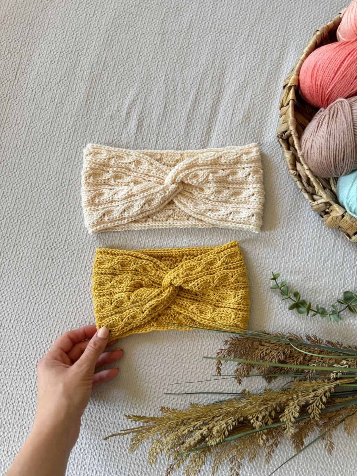 Two twisted crochet headband patterns, one cream and one yellow, are laid out on a textured surface next to a basket of yarn, dried plants, and a hand touching the yellow headband.