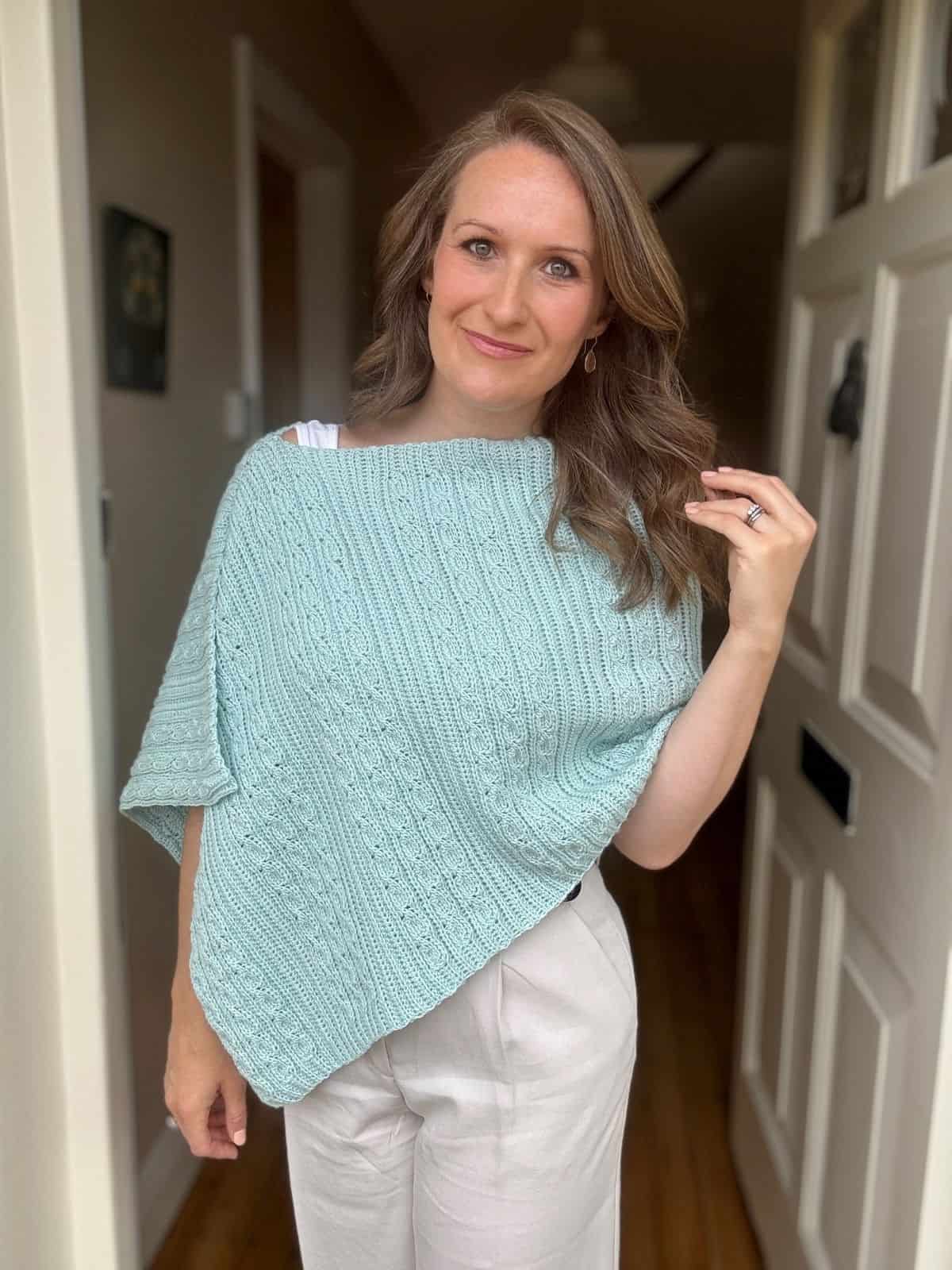 A woman stands indoors wearing a light blue crocheted poncho and light-colored pants, with her hand resting on her hair.