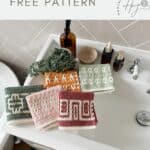 Five crocheted washcloths with geometric crochet washcloth patterns in green, pink, orange, and red are displayed on a white bathroom sink next to toiletries and a small plant.