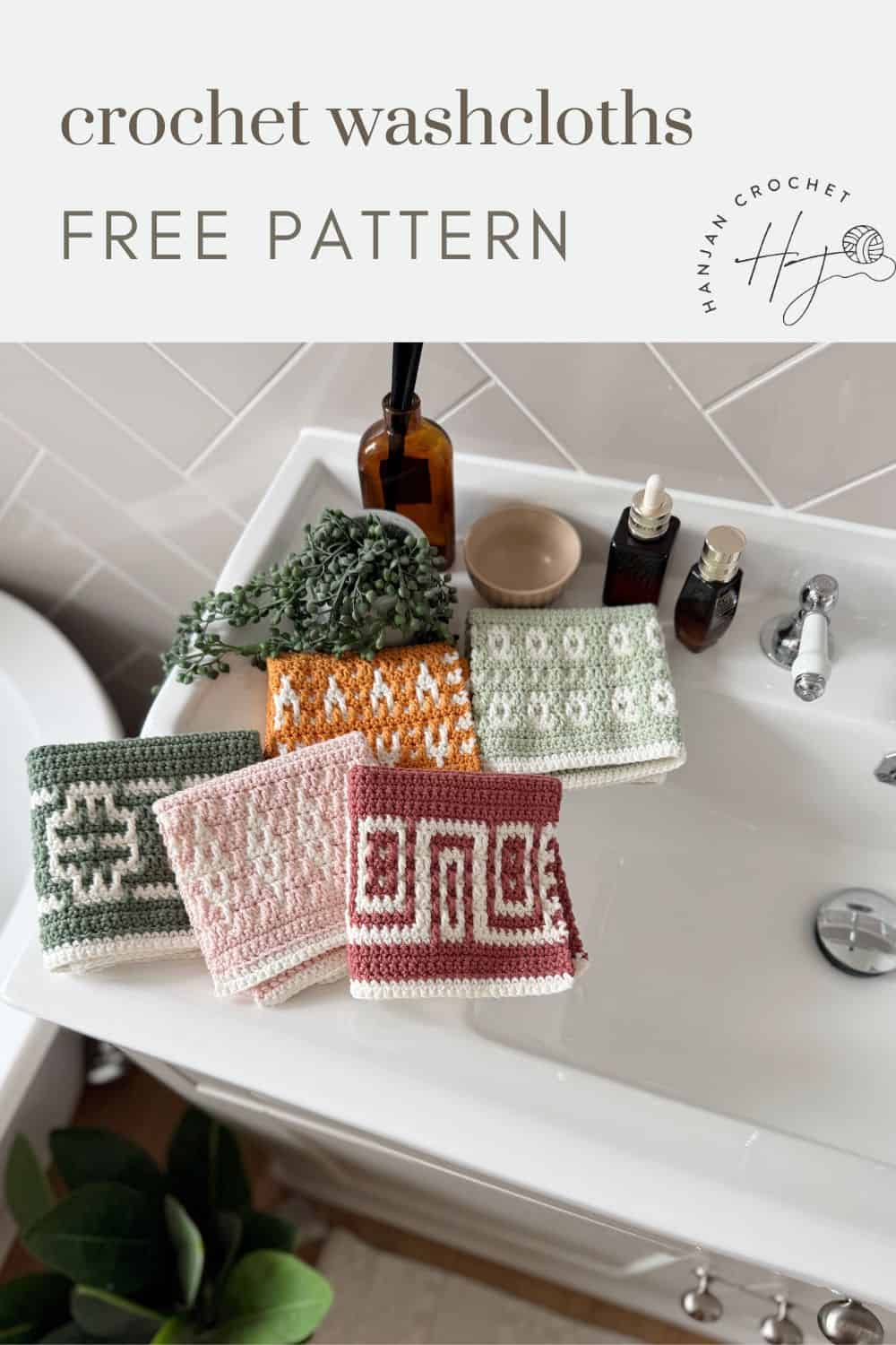 Five crocheted washcloths with geometric crochet washcloth patterns in green, pink, orange, and red are displayed on a white bathroom sink next to toiletries and a small plant.
