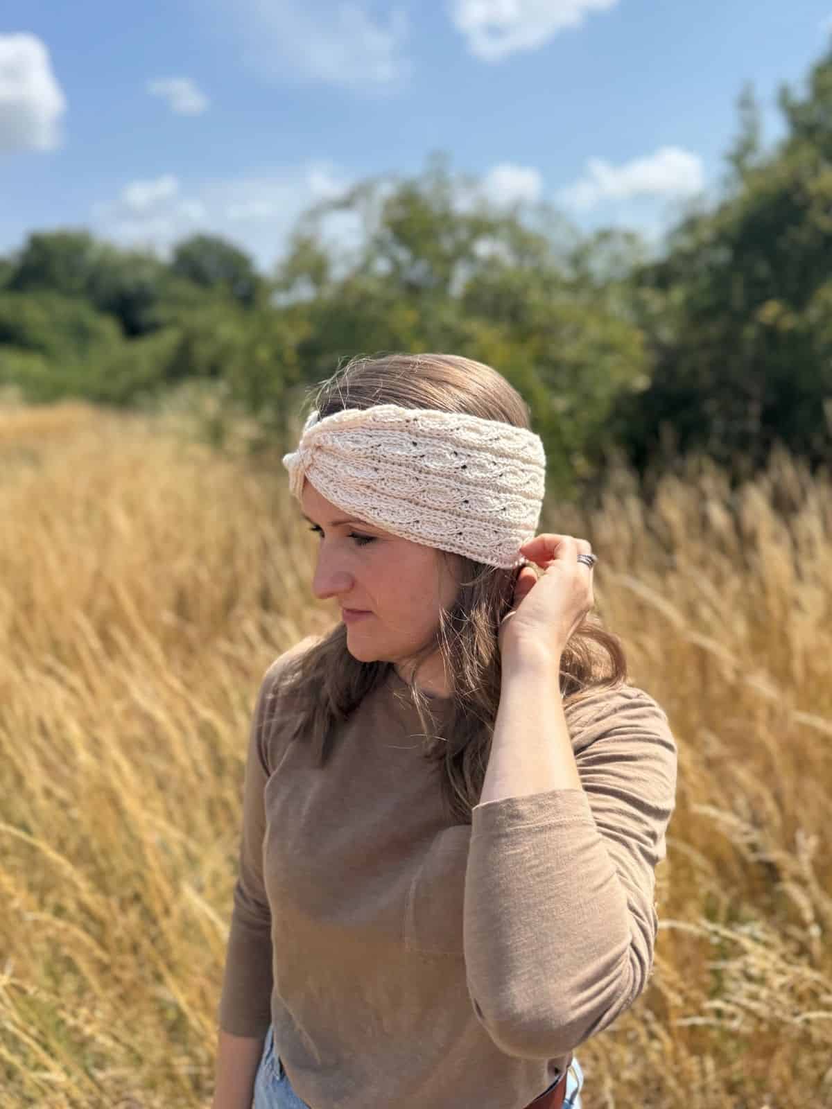 A woman wearing a twisted crochet headband pattern and a long-sleeve shirt stands in a grassy field with trees and blue sky in the background.