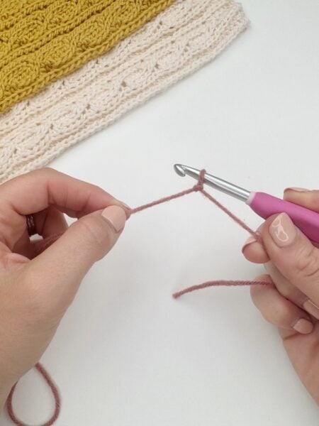 Hands holding a crochet hook and pink yarn, showing how to make a slip knot crochet for beginners; completed yellow and cream crocheted fabric appears in the background.