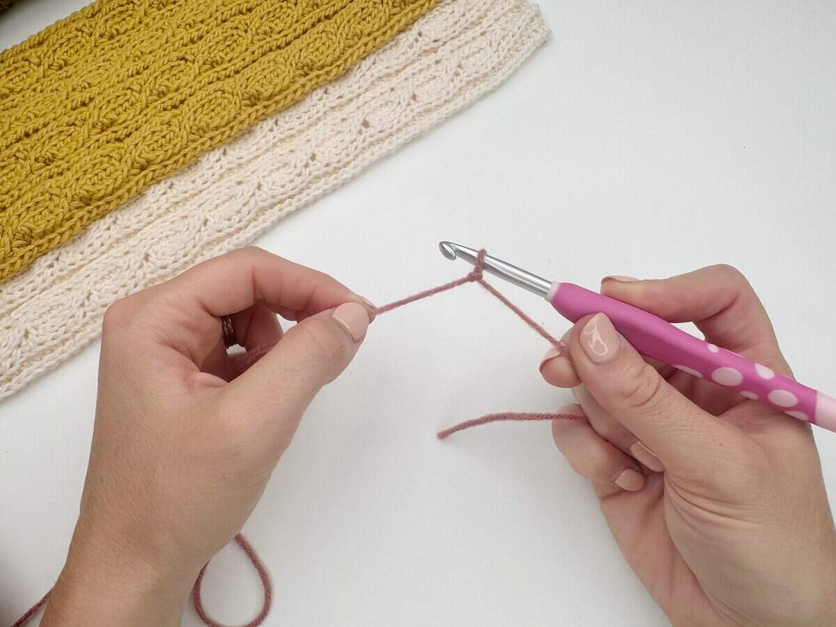 Hands holding a crochet hook and pink yarn, showing how to make a slip knot crochet for beginners; completed yellow and cream crocheted fabric appears in the background.