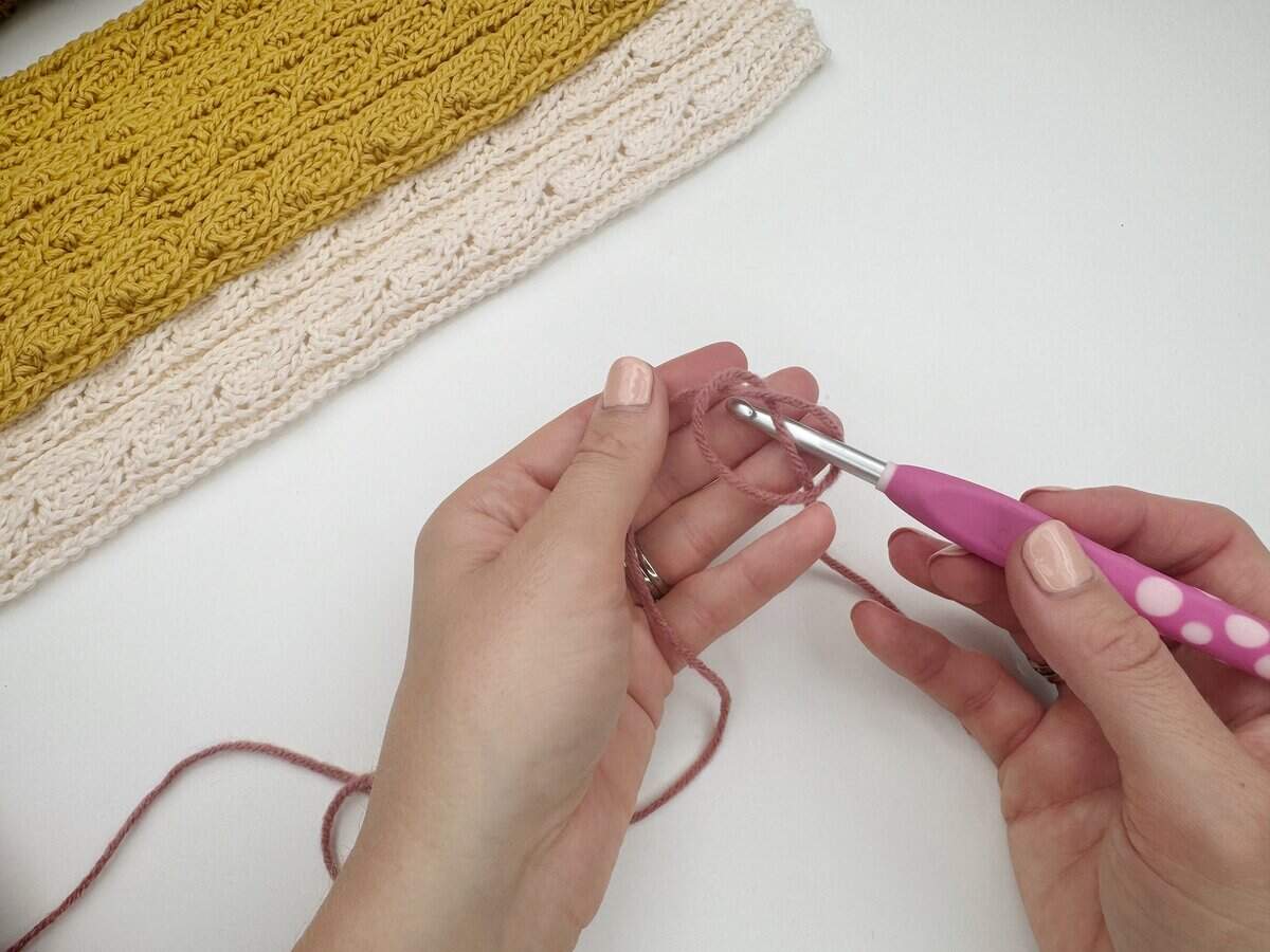 Hands holding yarn and a crochet hook, demonstrating how to make a slip knot crochet for beginners and start a new project with a chain; a finished yellow and cream crochet piece is in the background.