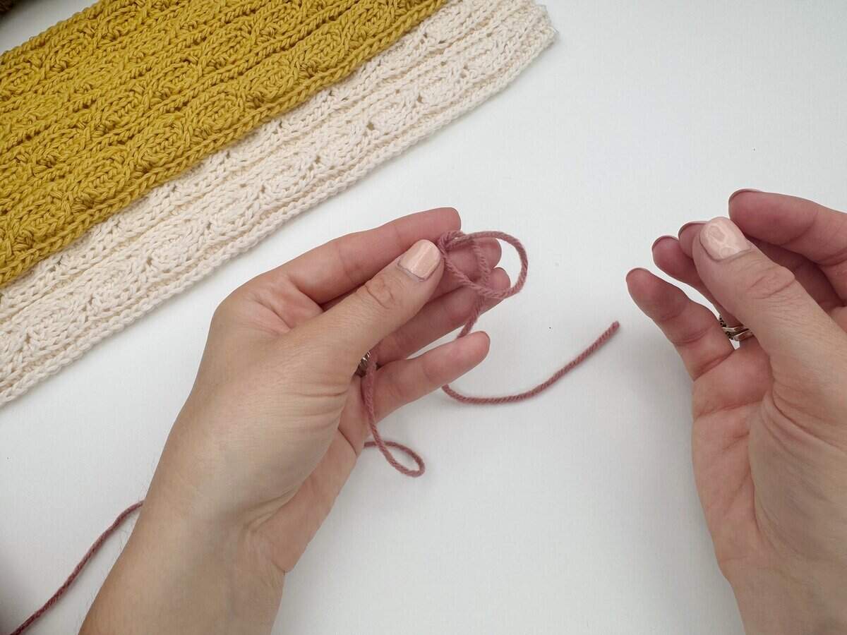 Two hands holding pink yarn, preparing to crochet, demonstrate how to make a slip knot crochet for beginners, with a finished yellow and white textured crochet piece in the background on a white surface.