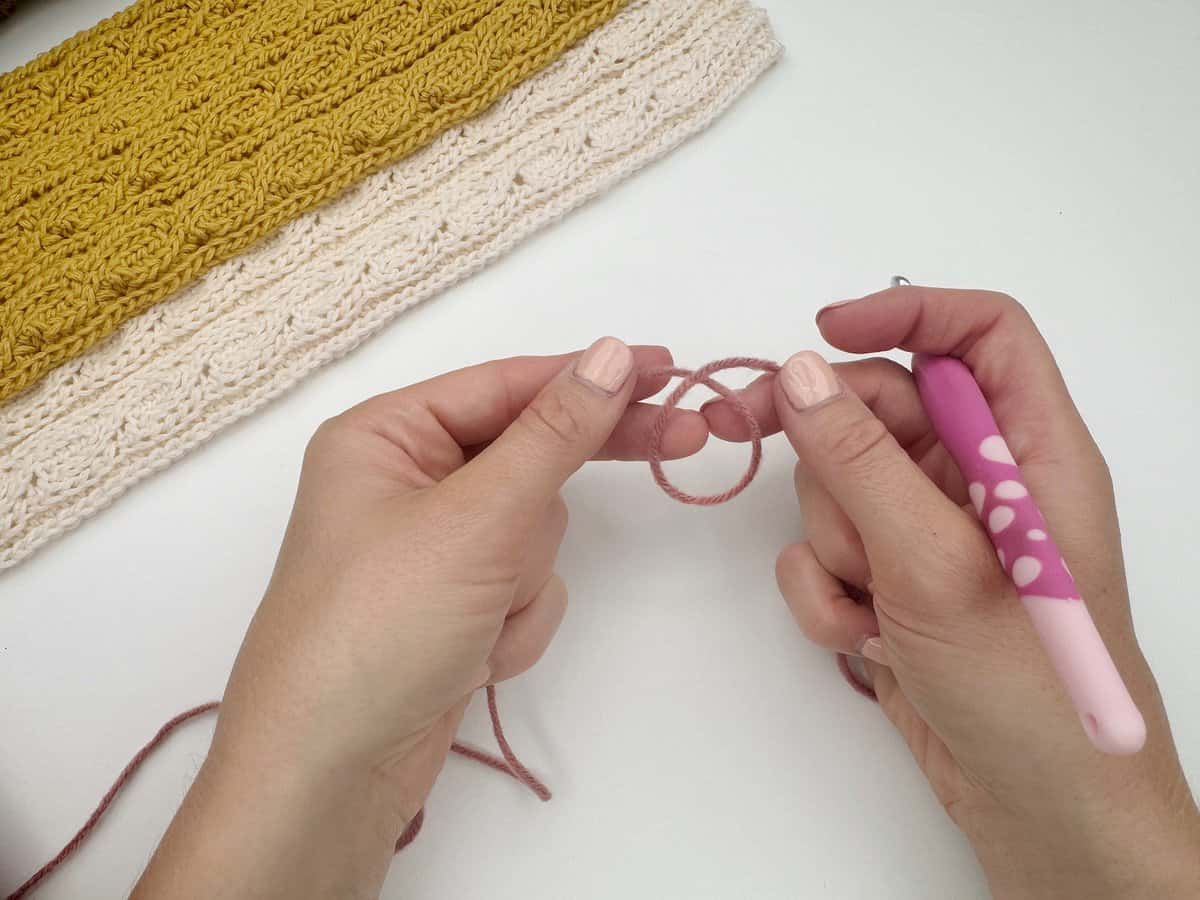 Two hands hold a pink yarn loop and a crochet hook above a white surface with a yellow and cream crocheted piece, perfect for showing how to make a slip knot crochet for beginners.