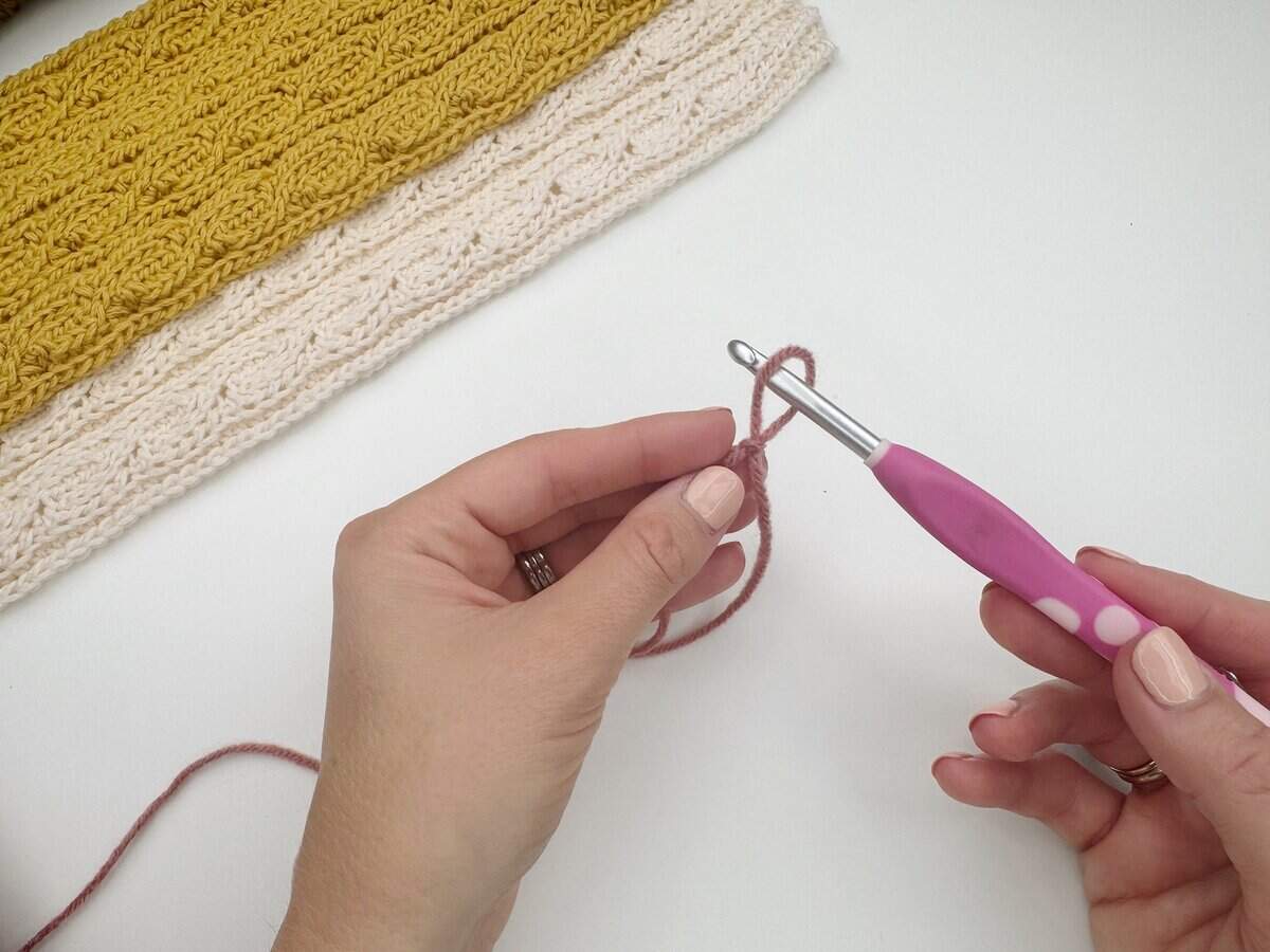 A person holds a pink crochet hook and yarn, preparing to crochet and learn how to make a slip knot crochet for beginners, with a folded yellow and cream crochet piece in the background.
