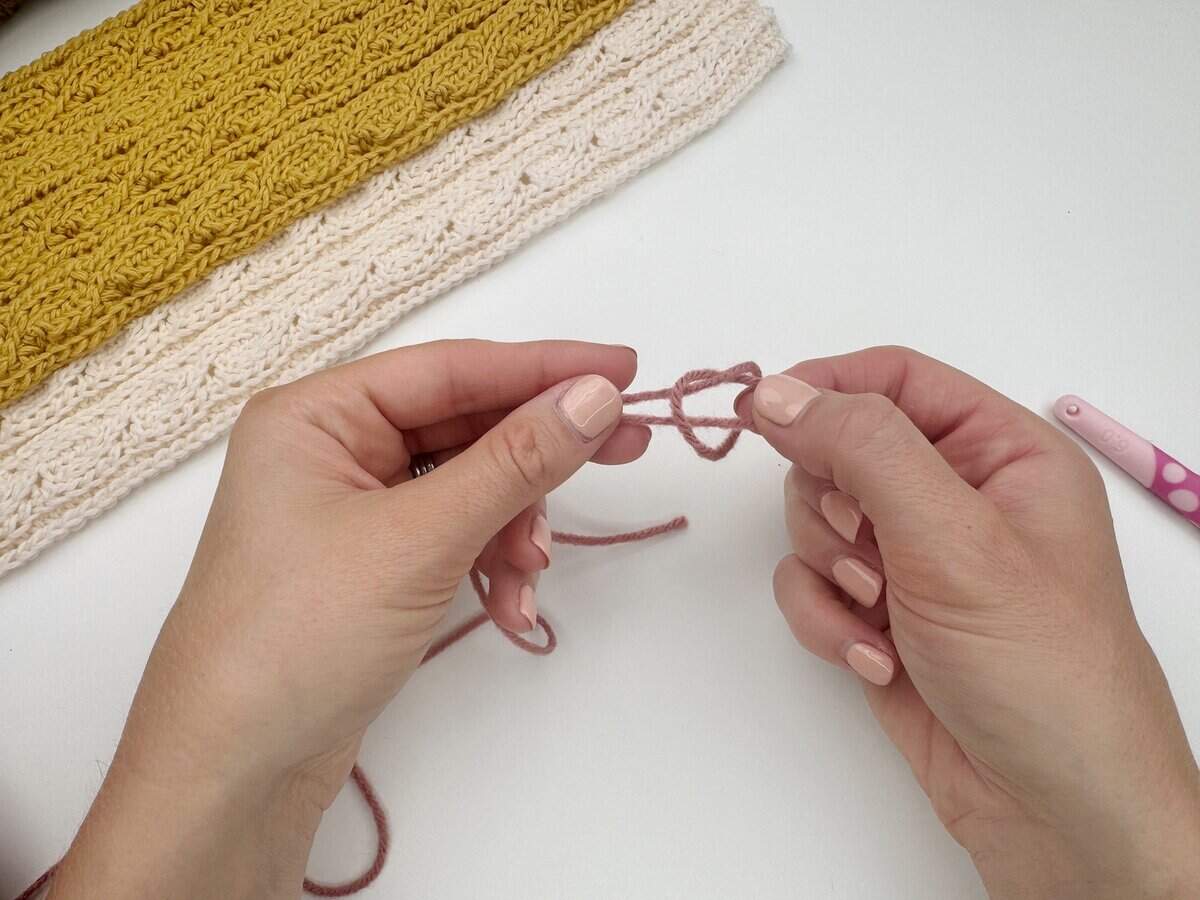 Hands demonstrating how to make a slip knot crochet for beginners with pink yarn, alongside a crochet hook and finished white and yellow crochet fabric pieces on a white surface.