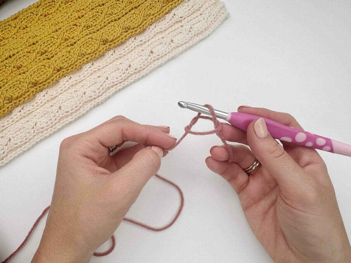 A person holds a crochet hook and pink yarn, starting a new crochet project—perfect for learning how to make a slip knot crochet for beginners. Above, a finished piece with yellow and cream stitches lies on the table.