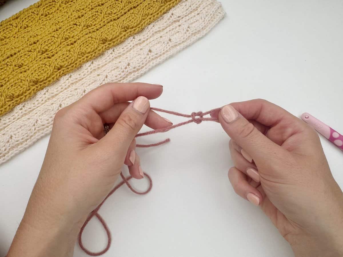 Close-up of hands demonstrating how to make a slip knot crochet for beginners with pink yarn, alongside a crocheted yellow and white piece and a pink crochet hook on a white surface.