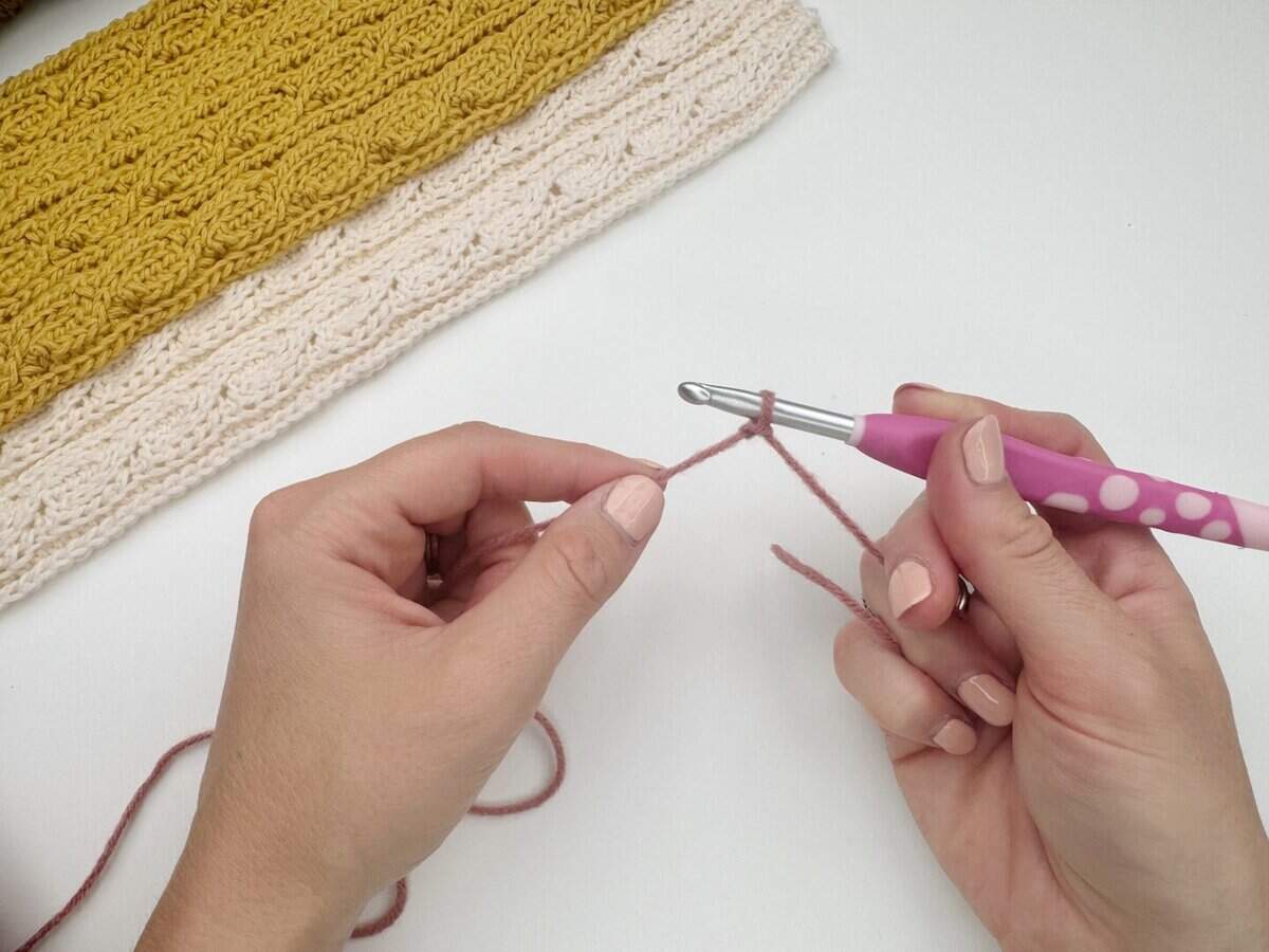 Hands holding a crochet hook and pink yarn, beginning a crochet chain—perfect for showing how to make a slip knot crochet for beginners. Folded yellow and cream crochet pieces rest in the background on a white surface.