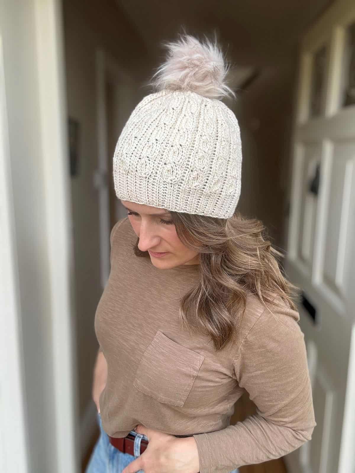 A woman wearing a beige crochet hat with a pom-pom, crafted in a short row crochet hat pattern, and a brown long-sleeve shirt stands indoors with her hands in her pockets, looking down.