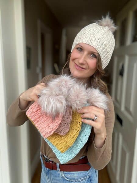 Woman in a beige crochet hat holds several colorful short row crochet hat pattern designs, each topped with a fluffy pom-pom, standing indoors near a white door.