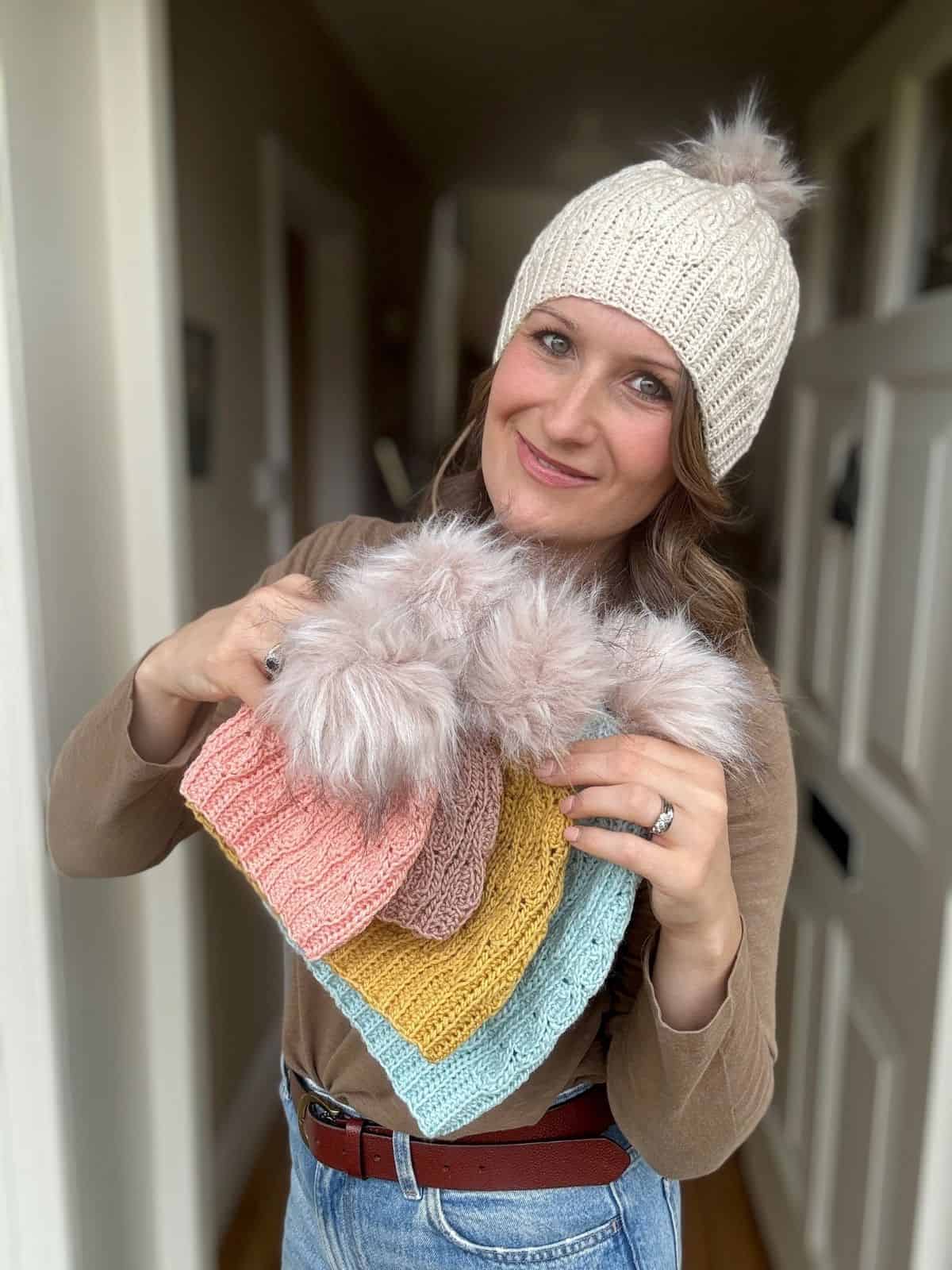 Woman in a beige crochet hat holds several colorful short row crochet hat pattern designs, each topped with a fluffy pom-pom, standing indoors near a white door.