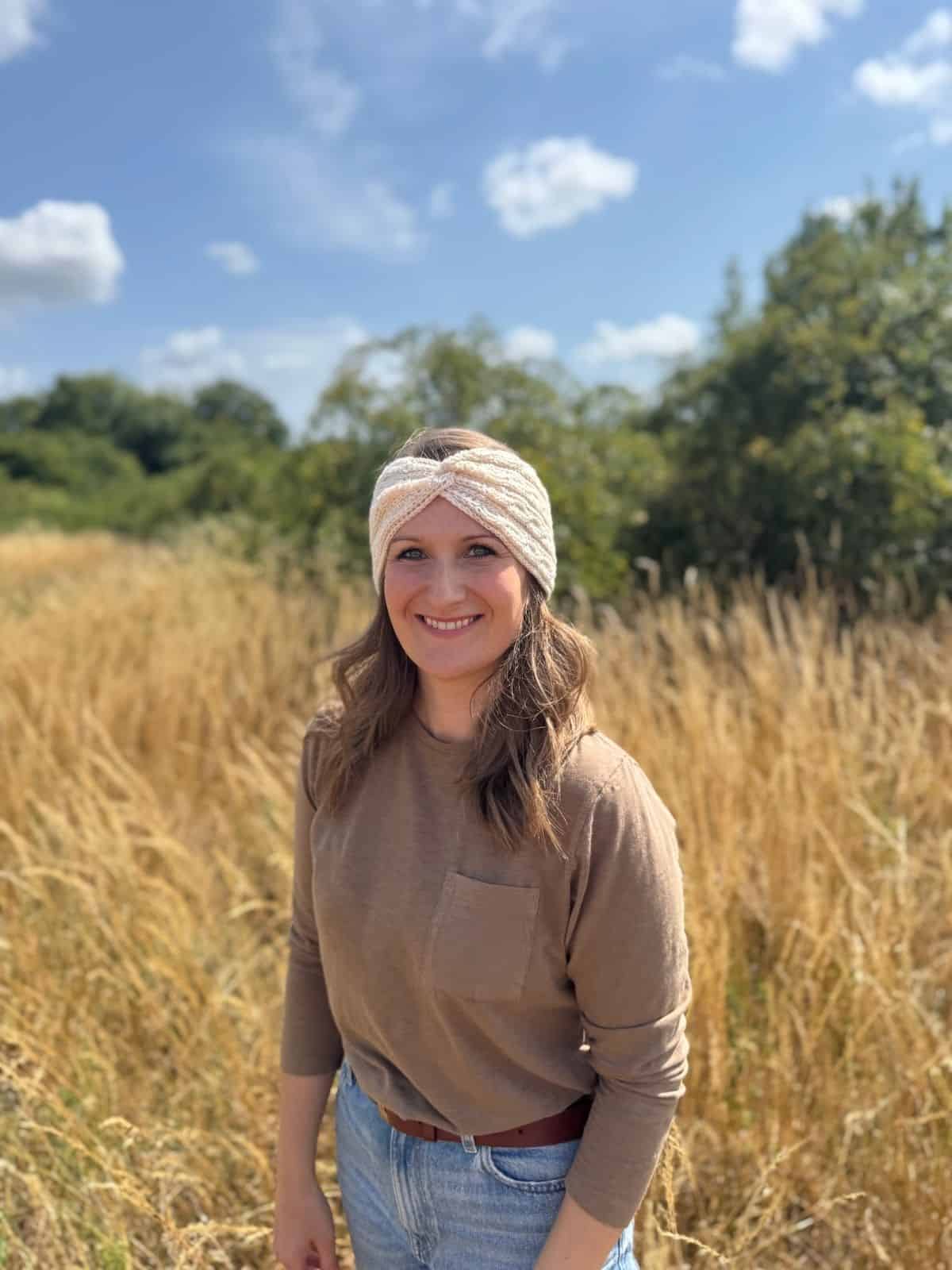 A woman wearing a twisted crochet headband pattern and a brown long-sleeve shirt stands smiling in a sunny, grassy field with trees in the background.