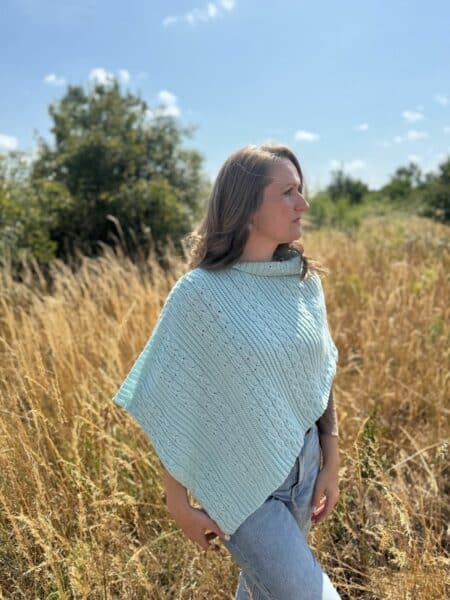 A woman wearing a light blue crochet poncho stands in a field of tall dry grass under a clear sky, looking to the side.