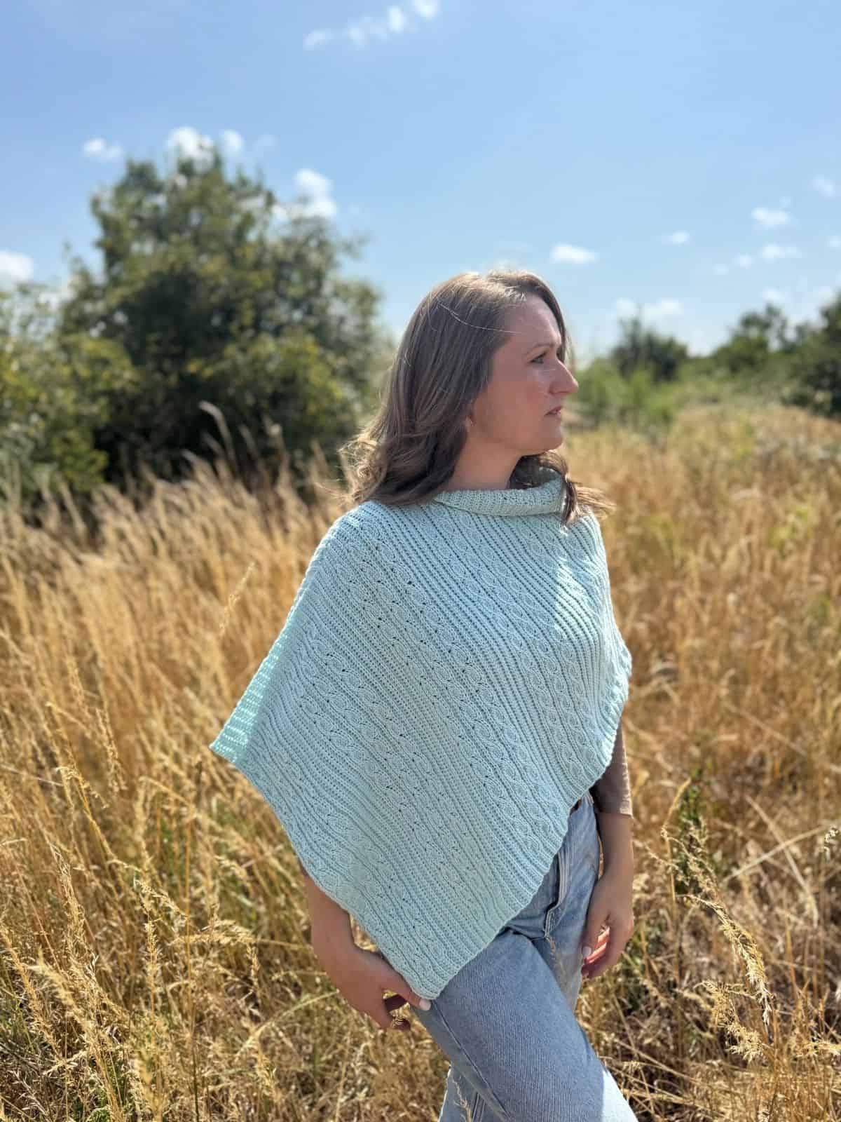 A woman wearing a light blue crochet poncho stands in a field of tall dry grass under a clear sky, looking to the side.