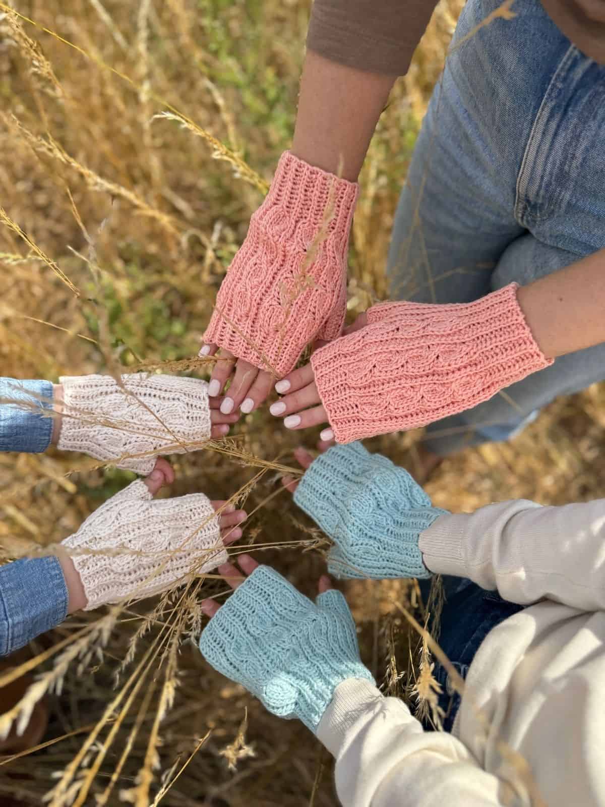 Five people wearing pastel crochet fingerless mittens, inspired by a free pattern, stand in a circle outdoors and extend their hands toward the center over dry grass.