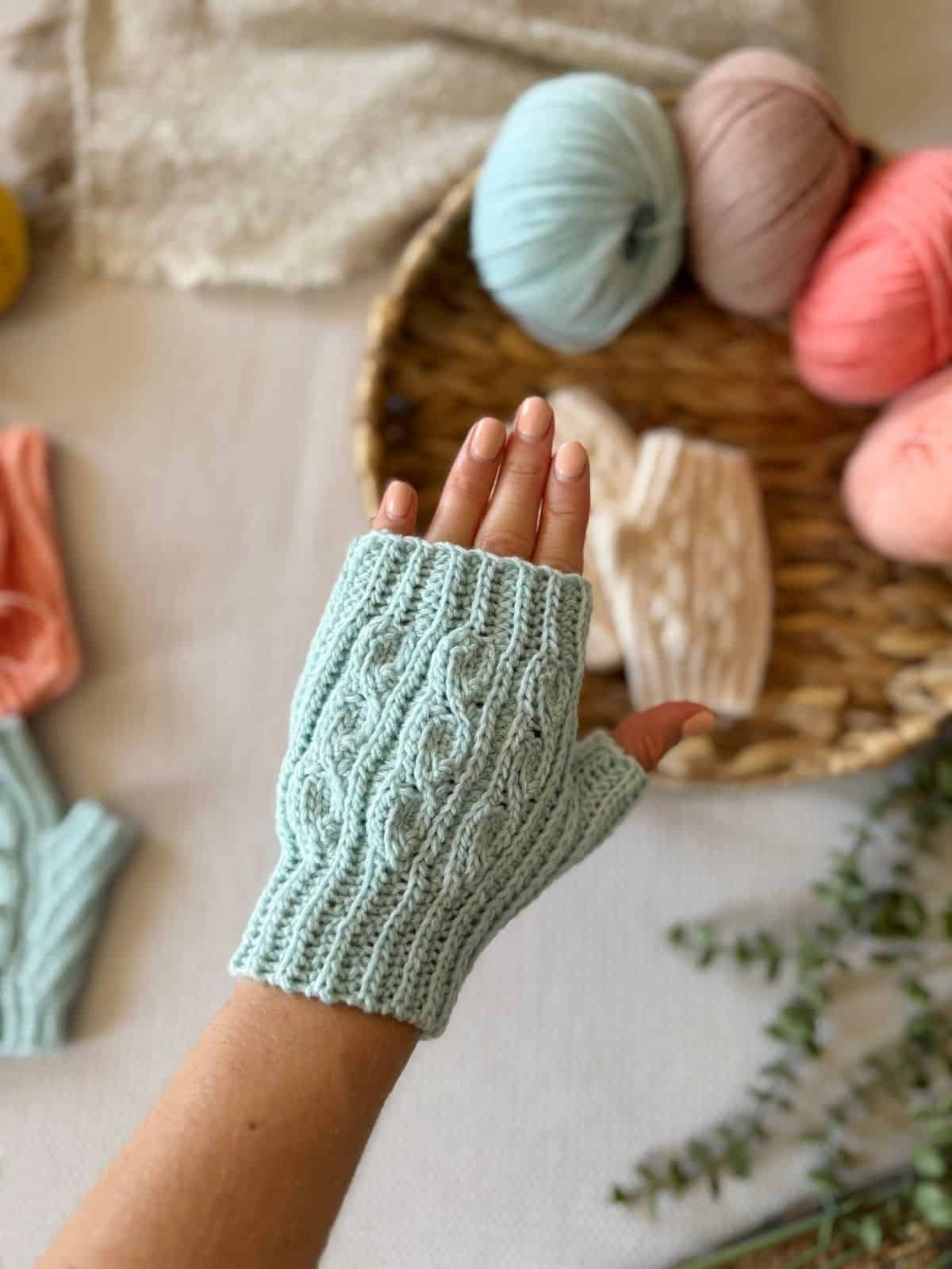 A hand wearing a light blue crocheted fingerless glove is held up, with yarn balls and more crocheted items in the background—perfect inspiration if you're searching for a crochet fingerless mittens pattern free of charge.