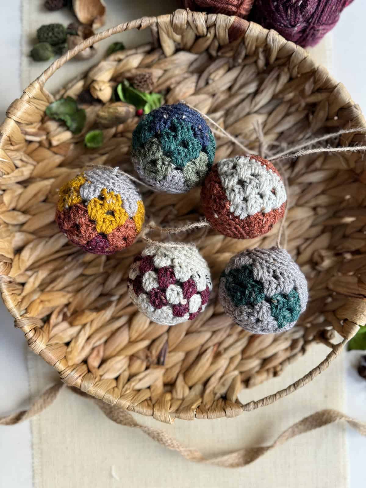 A woven basket contains five colorful, hand-crocheted baubles made from easy Christmas crochet patterns, each featuring unique designs and a string loop for hanging.